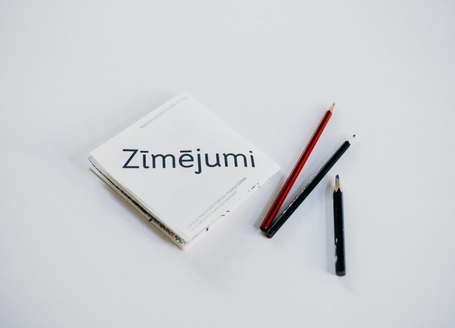 Balta grāmata ar uzrakstu Zīmējumi un trīs zīmuļi uz gaišax fona.