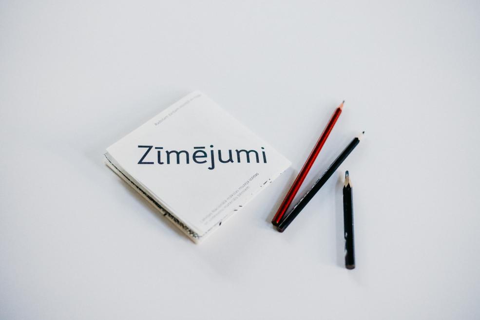 Balta bukleta vāka ar uzrakstu Zīmējumi blakus trim zīmuļiem uz gaiša fona.