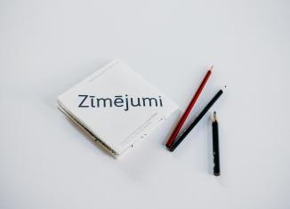 Balta grāmata ar uzrakstu Zīmējumi un trīs zīmuļi uz gaišax fona.