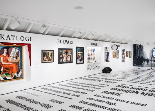 Muzeja ekspozīcijas zāle ar gleznām un fotogrāfijām pie sienas zem uzrakstiem “Skatlogi”, “Bulvāri” un “Kioski”, bet uz grīdas izvietoti liela formāta teksti.