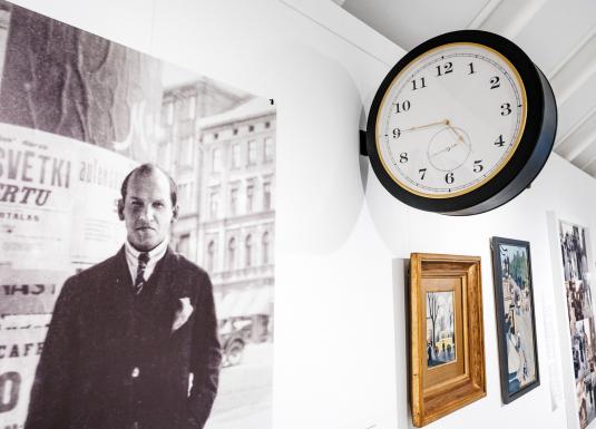 Muzeja ekspozīcijā pie sienas redzama vēsturiska fotogrāfija ar vīrieti pilsētas vidē, blakus izvietotas gleznas un apaļš sienas pulkstenis.