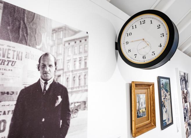 Muzeja ekspozīcijā pie sienas redzama vēsturiska fotogrāfija ar vīrieti pilsētas vidē, blakus izvietotas gleznas un apaļš sienas pulkstenis.