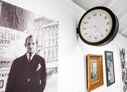 Muzeja ekspozīcijā pie sienas redzama vēsturiska fotogrāfija ar vīrieti pilsētas vidē, blakus izvietotas gleznas un apaļš sienas pulkstenis.