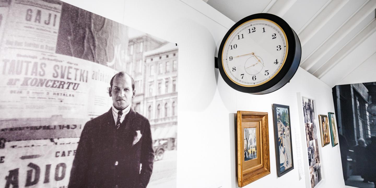 Muzeja ekspozīcijā pie sienas redzama vēsturiska fotogrāfija ar vīrieti pilsētas vidē, blakus izvietotas gleznas un apaļš sienas pulkstenis.