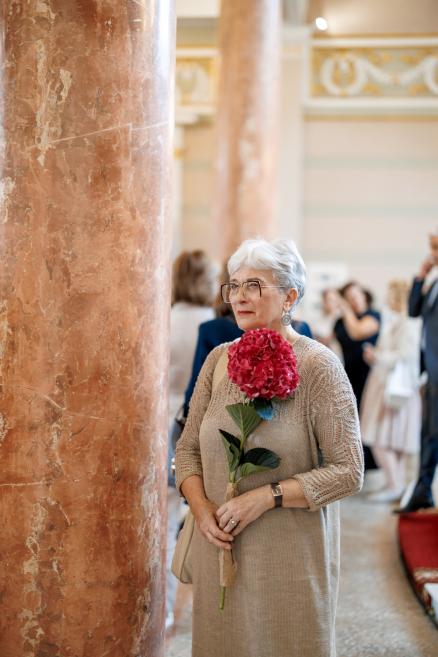 A woman with gray hair and glasses holds a large pink hydrangea in the Latvian National Museum of Art hall.