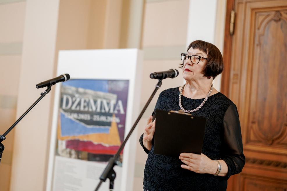 A woman in black with a folder speaks at the microphone during the “Džemma” exhibition opening.