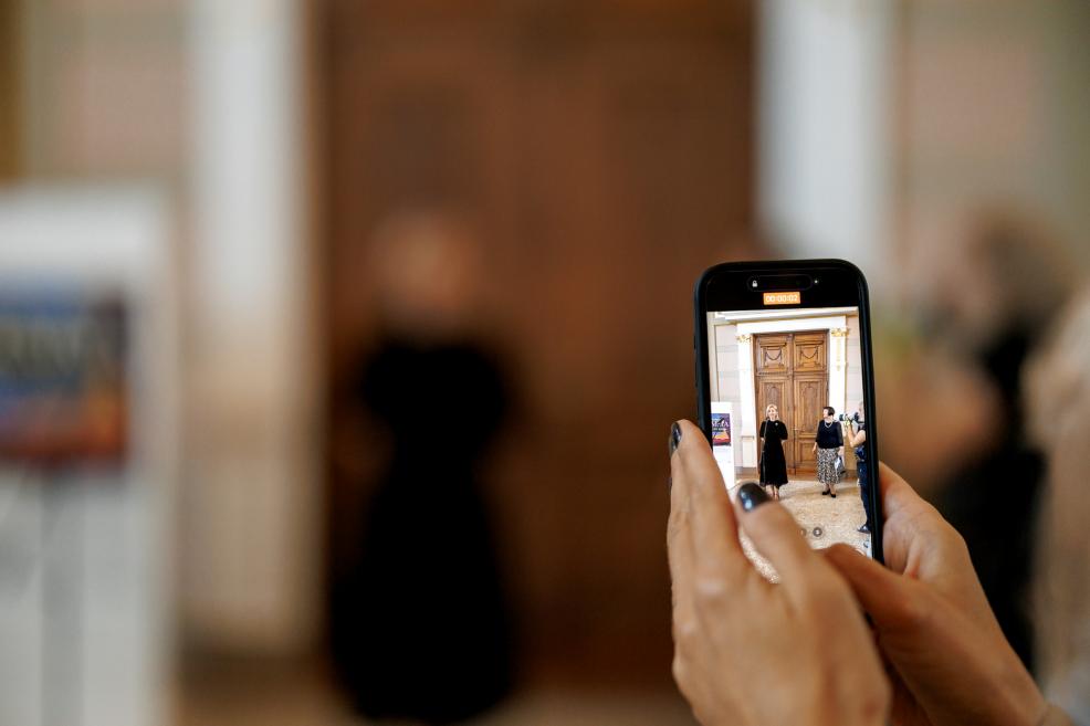 A phone held in hands shows a woman speaking at the exhibition opening.