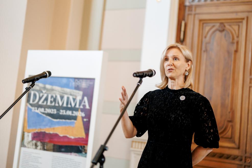A woman in a black dress speaks at the microphone with the “Džemma” exhibition poster in the background.
