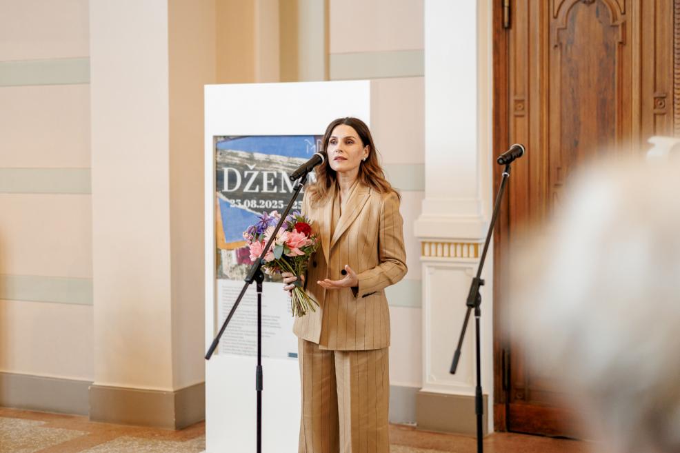 A woman in a light suit holding a bouquet speaks into a microphone at the opening of the exhibition “Džemma.”