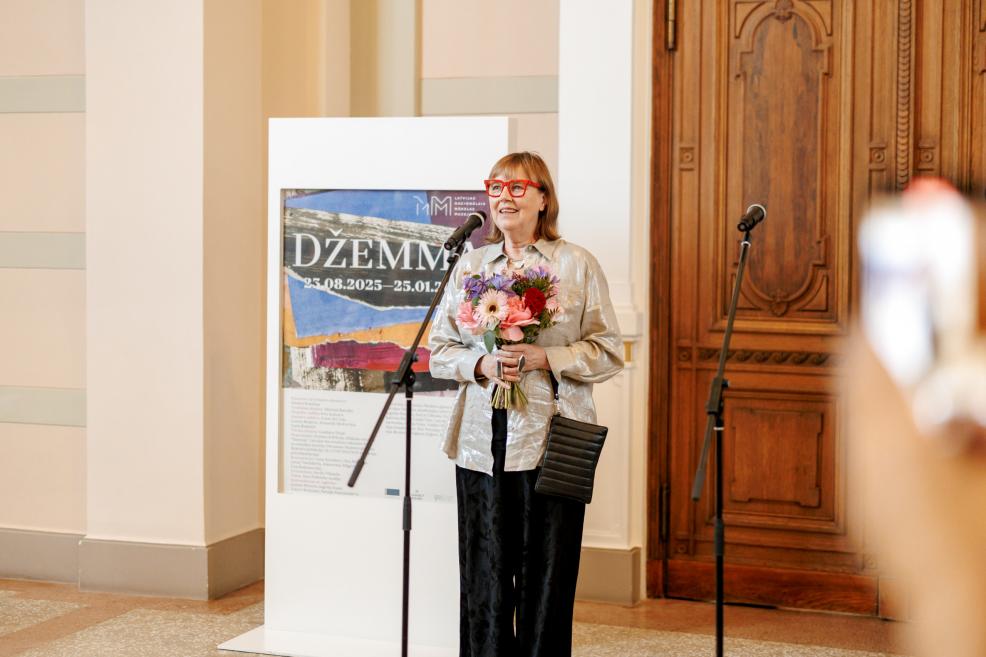 A woman with bright red glasses and a bouquet speaks into a microphone at the “Džemma” exhibition.