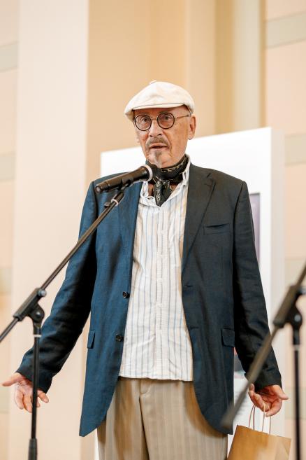 A man in a white cap and dark jacket speaks into a microphone at the opening of the “Džemma” exhibition.