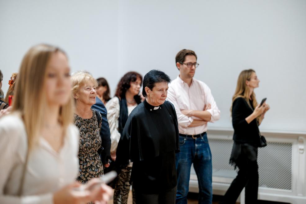 Visitors stand in a white exhibition hall attentively listening to a speech.