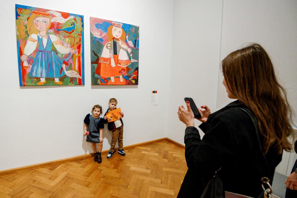Two children pose in front of colorful paintings while a woman takes their photo.