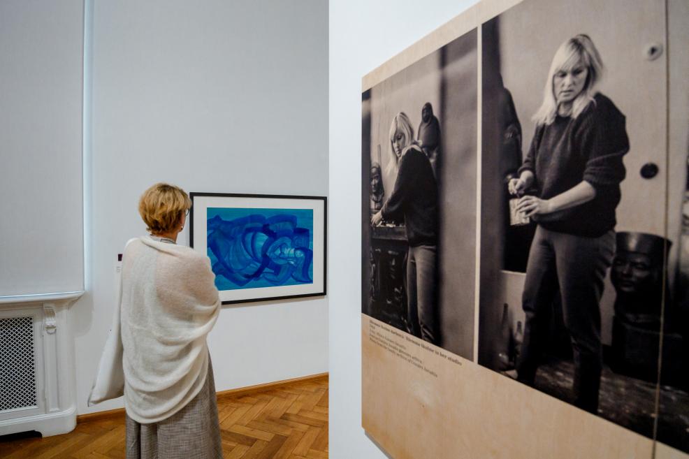 A woman views a work by Džemma Skulme, next to a large black-and-white photo of the artist.