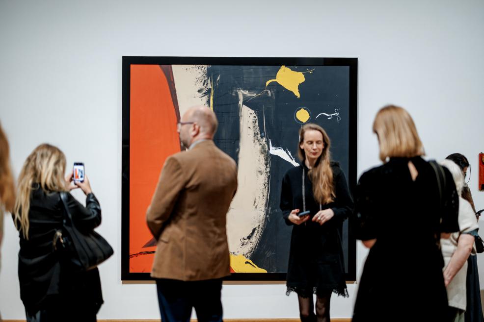 A woman with long hair in a black dress stands near a large abstract painting with red, black, and yellow forms.