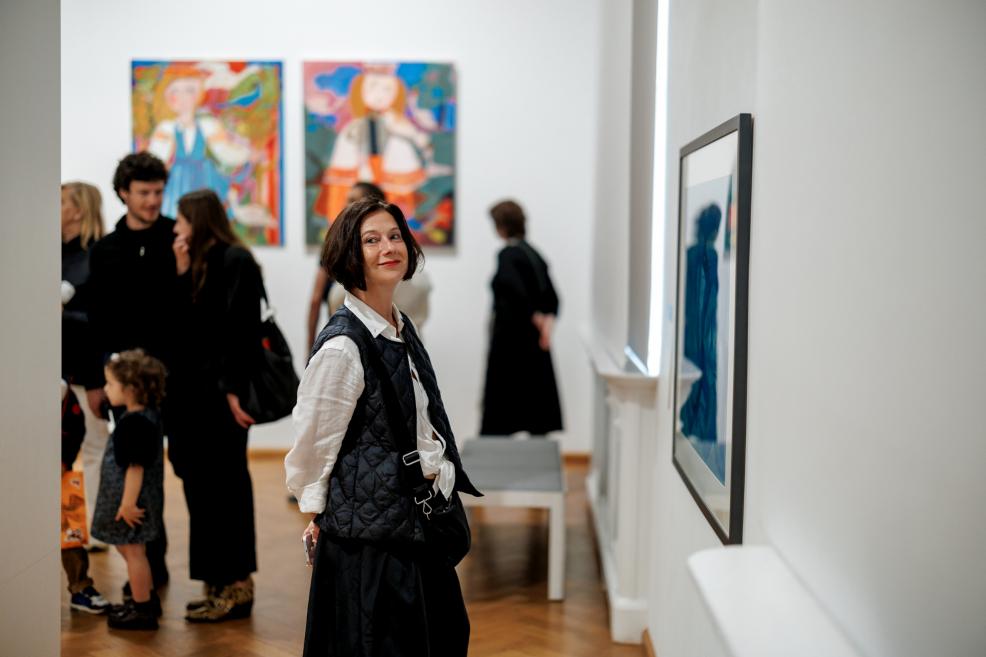 A woman in a white blouse and black vest smiles while standing in the exhibition hall with colorful paintings in the background.