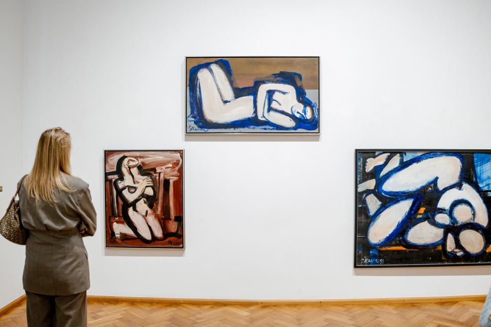 A woman in a gray suit looks at three paintings depicting human figures in blue and red tones.