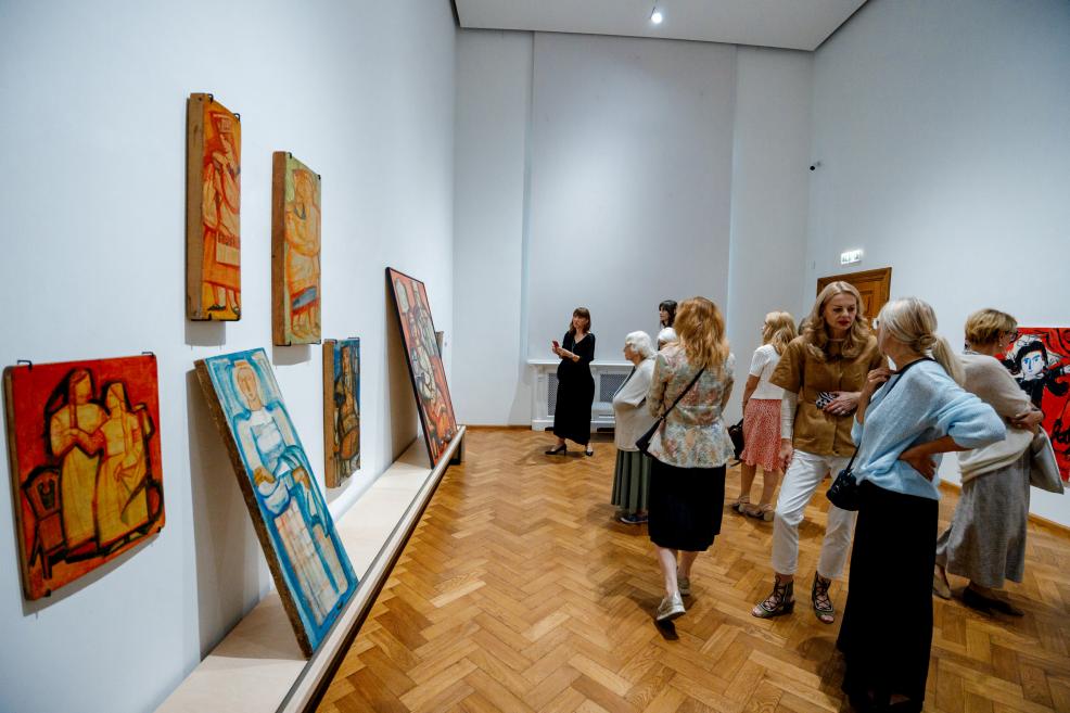 Exhibition visitors view several paintings with stylized figures and bright colors displayed on panels.