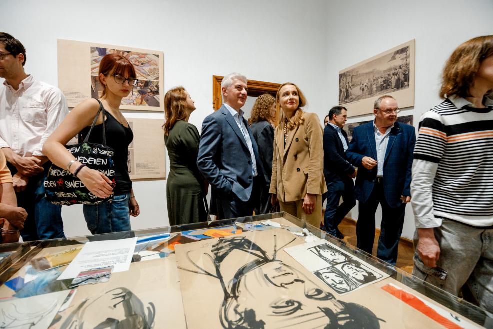 A group of people stand around a display case with drawings and graphic works, viewing the exhibition.