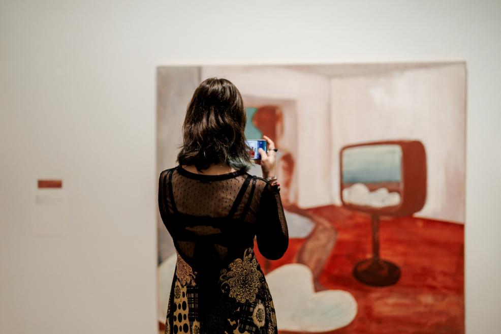 A woman in a black dress photographs a painting of an interior with a television on a reddish-brown floor.