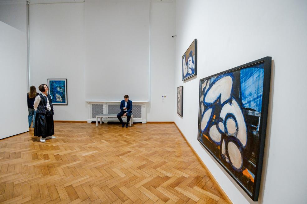 In the exhibition hall, a man sits on a bench while a woman views large abstract paintings in blue and white colors.