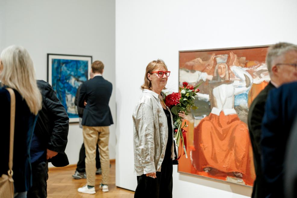 A woman with red glasses holding red flowers stands beside a large painting of a female figure in an orange dress.