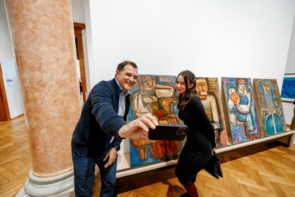 A man and a woman smile while taking a selfie in front of paintings with stylized human figures.