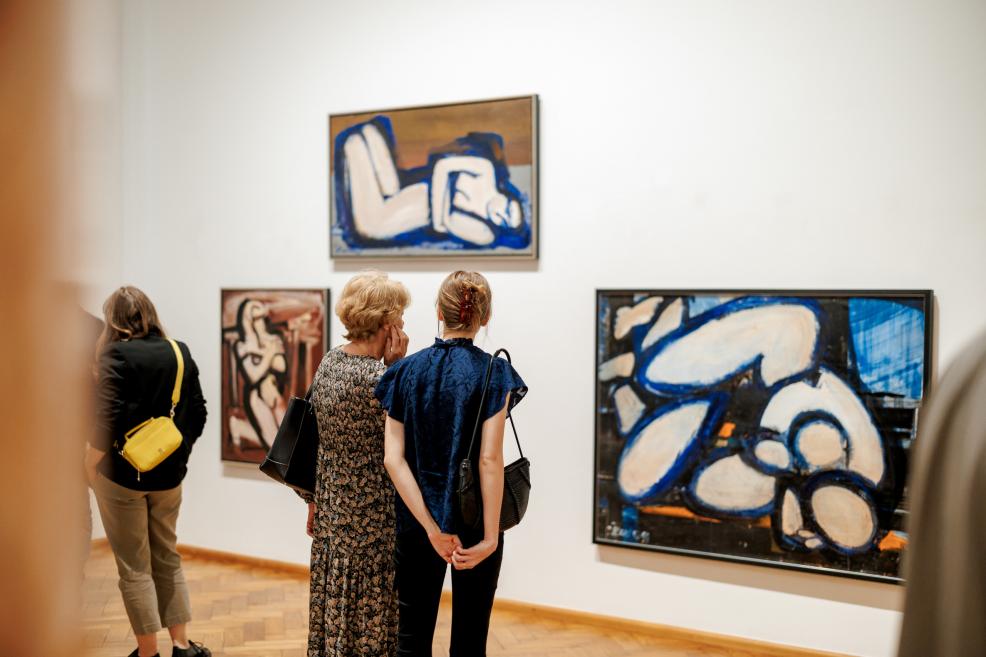 Two women observe paintings of abstract human figures in blue and red tones.