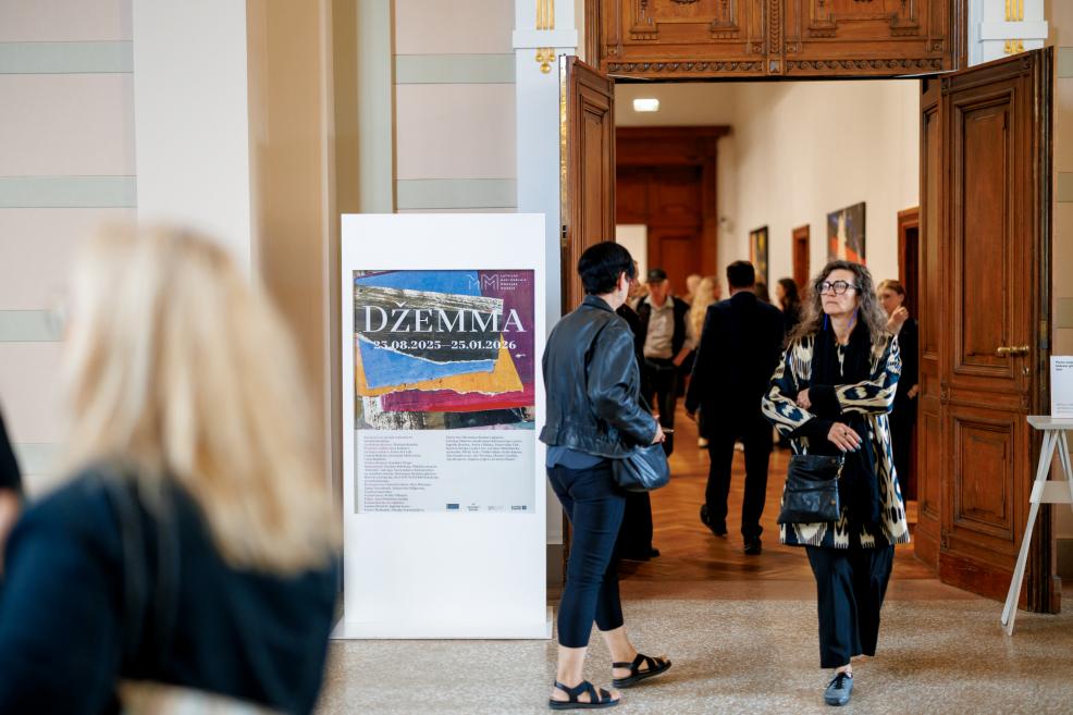 In the museum foyer, a poster for the exhibition “Džemma” is displayed with visitors in the background.