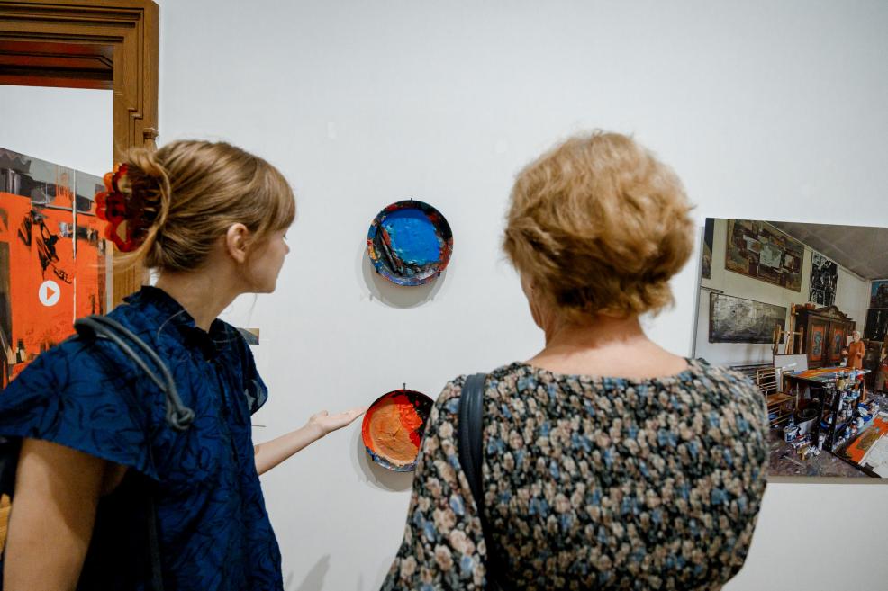 Two women view colorful paint palettes and a studio photograph displayed on the wall.