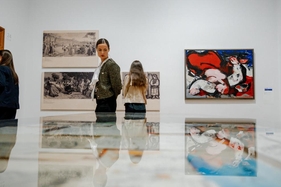 Women stand near paintings and large-scale graphic reproductions in the exhibition hall.