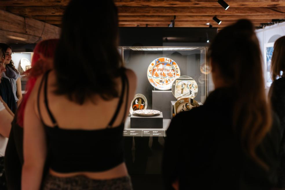 Apmeklētāji muzeja zālē skatās uz vitrīnā izstādītiem dekoratīviem keramikas šķīvjiem ar krāsainiem ornamentiem.