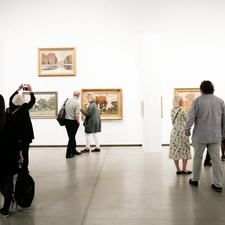 Muzeja apmeklētāji apskata gleznas izstāžu zālē, vairāki no viņiem fotografē eksponātus.
