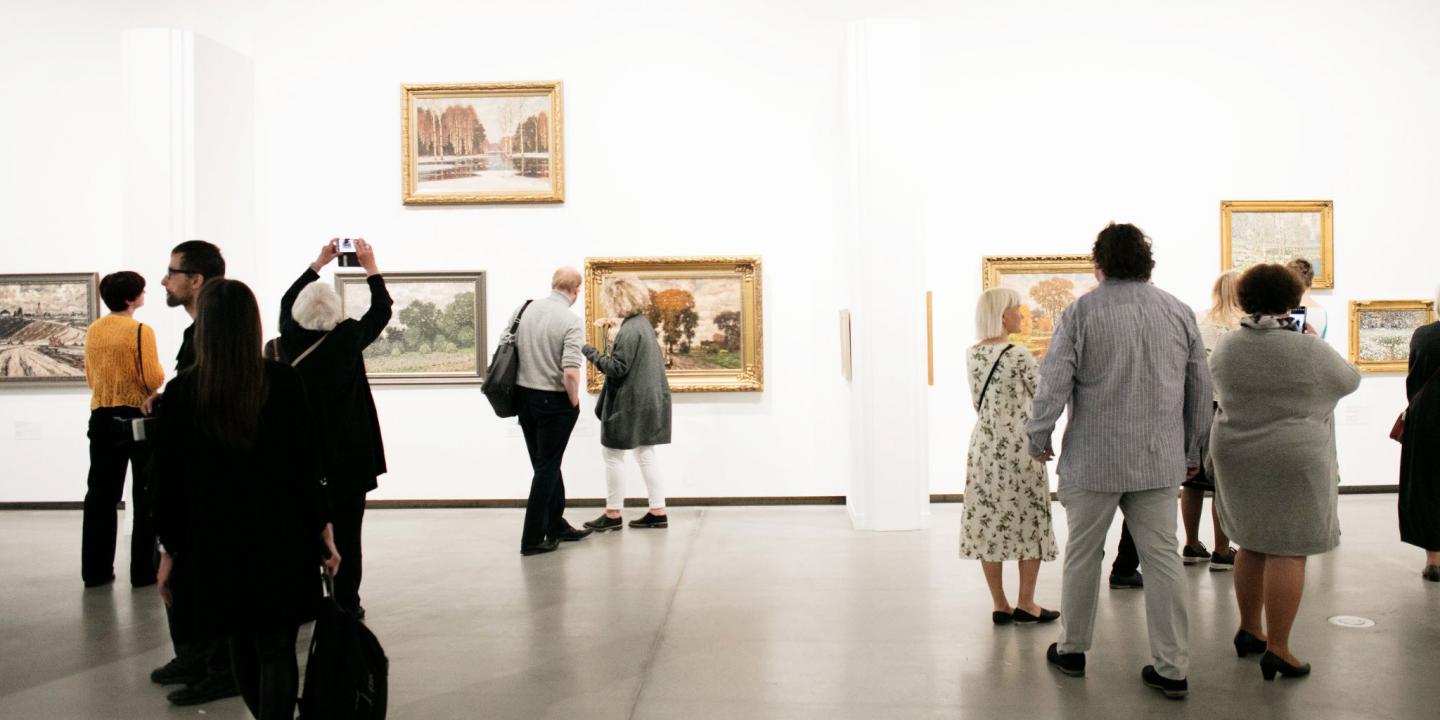 Muzeja apmeklētāji apskata gleznas izstāžu zālē, vairāki no viņiem fotografē eksponātus.