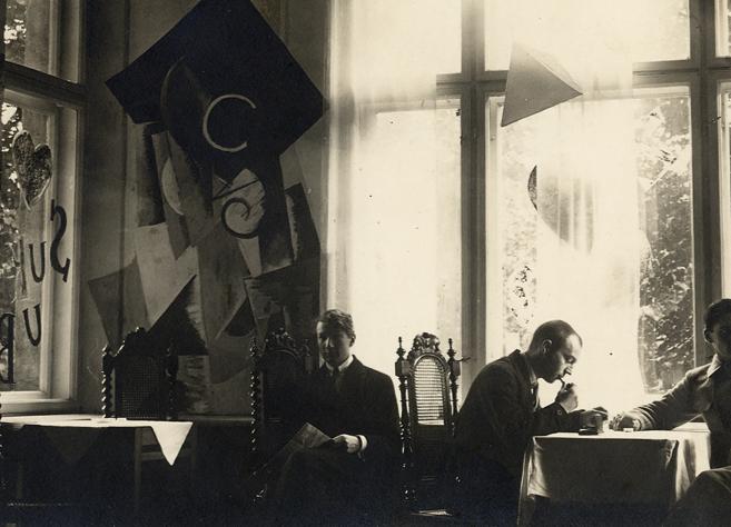 Melnbalta fotogrāfija ar vairākiem vīriešiem pie galdiņiem gaišā telpā ar lieliem logiem un dekoratīvām ģeometriskām formām pie sienām.