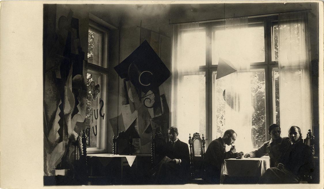 Melnbalta fotogrāfija ar četriem vīriešiem gaišā telpā pie logiem, kur redzami abstrakti papīra dekorējumi un galdi ar baltiem galdautiem.