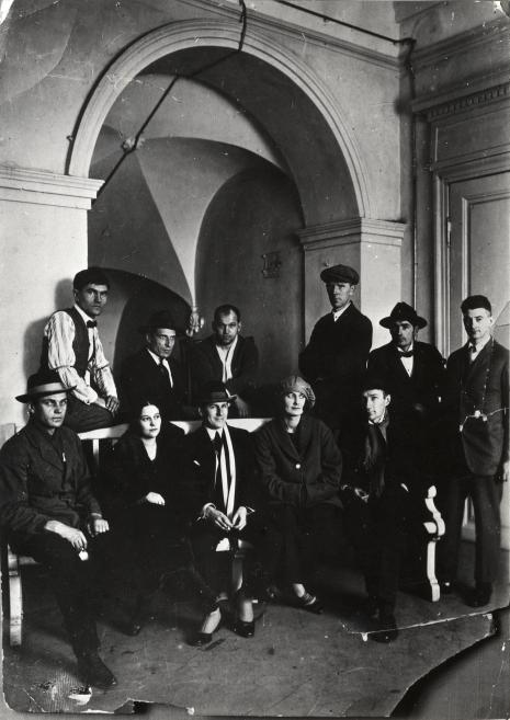 Melnbalta grupas fotogrāfija, kurā astoņi vīrieši un divas sievietes pozē telpā ar lielu arkveida nišu un durvīm fonā.