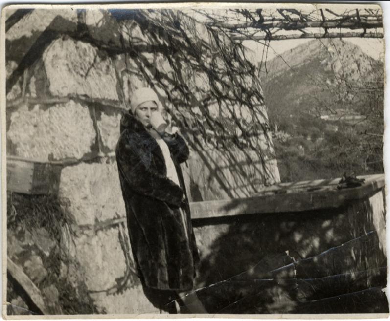 Melnbalta fotogrāfija ar sievieti mētelī un cepurē, kas stāv pie akmens sienas zem koku ēnām.