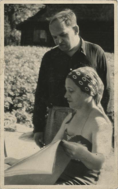 Black-and-white photograph of a man and a woman by a table outdoors, with the woman wearing a patterned headscarf and holding an open book or album.