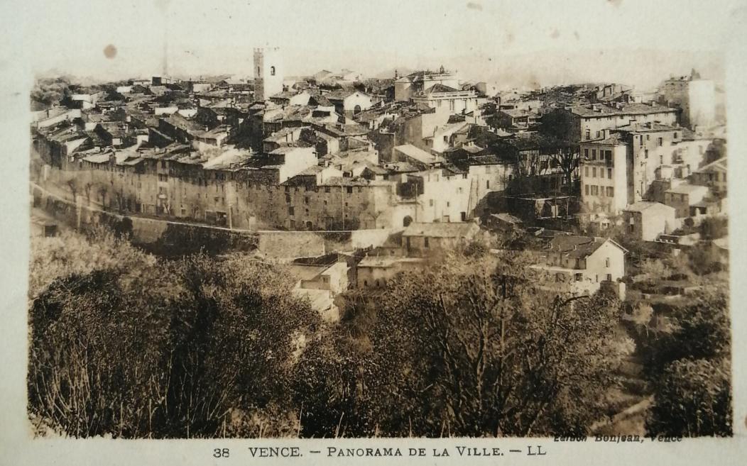 Sepijas tonī veidots Vansas pilsētas panorāmas skats ar blīvu apbūvi, sarkanīgiem jumtiem un kokiem priekšplānā.
