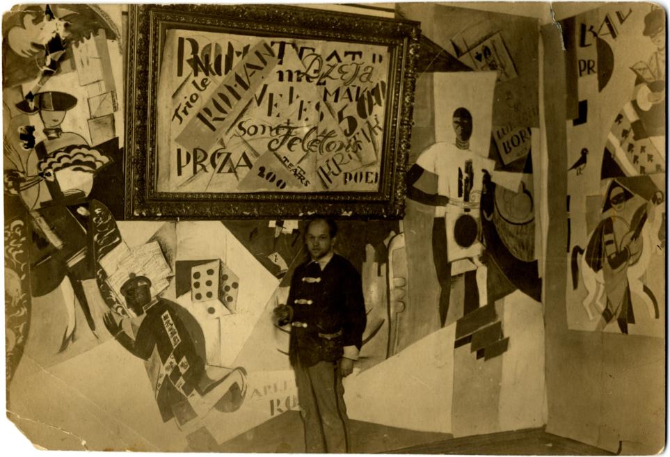 A man stands in an interior beside wall paintings and collages in an avant-garde style, including a large framed composition with text.