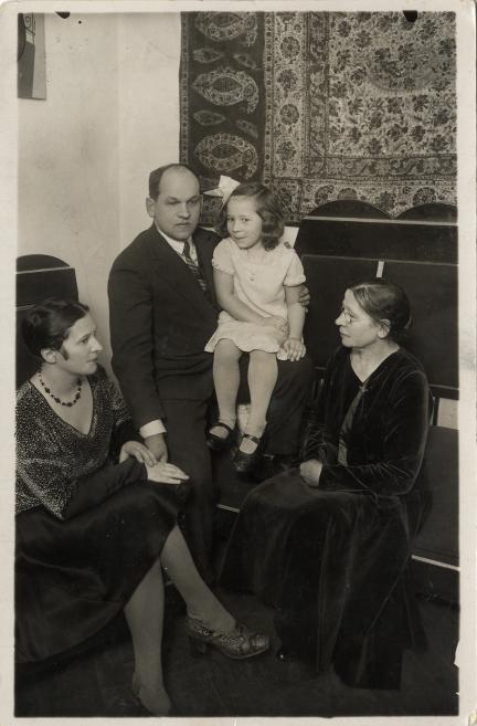 Melnbalta fotogrāfija ar vīrieti uz sola, kurš tur klēpī mazu meiteni ar baltu banti, bet abās pusēs sēž divas sievietes, fonā pie sienas redzams ornamentēts paklājs.