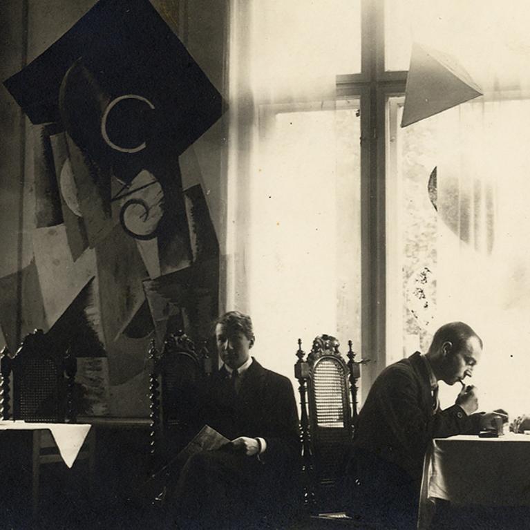 Melnbalta fotogrāfija ar vairākiem vīriešiem pie galdiņiem gaišā telpā ar lieliem logiem un dekoratīvām ģeometriskām formām pie sienām.