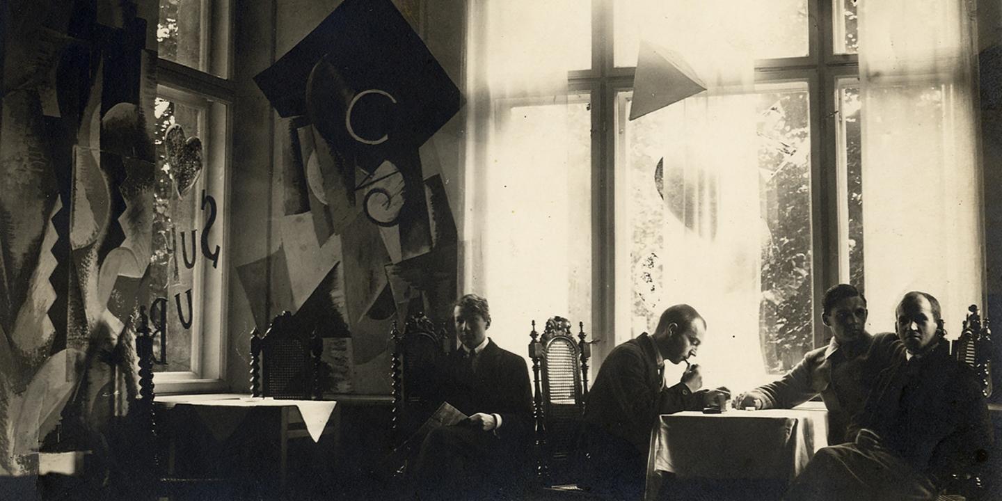 Melnbalta fotogrāfija ar vairākiem vīriešiem pie galdiņiem gaišā telpā ar lieliem logiem un dekoratīvām ģeometriskām formām pie sienām.