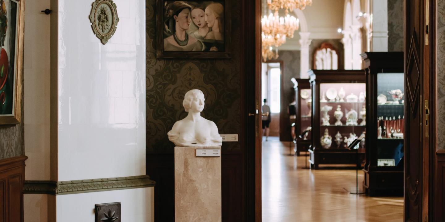 Vēsturiska muzeja interjers ar skulptūru uz postamenta, gleznām un izgaismotām vitrīnām fonā.