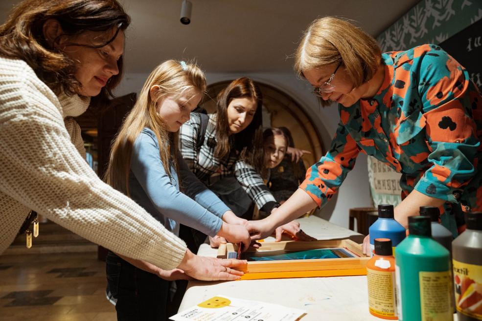 Pieaugušie un bērni kopā piedalās radošā sietspiedes darbnīcā, izmantojot krāsas un rāmjus.