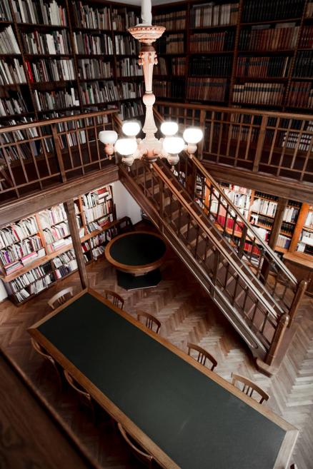 Latvijas Nacionālā mākslas muzeja bibliotēkas kopskats no augšas ar koka kāpnēm, lielu lasāmgalds un grāmatplauktiem gar sienām.