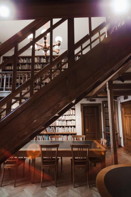 Latvijas Nacionālā mākslas muzeja bibliotēkas interjers ar koka kāpnēm, galdiem un grāmatplauktiem, kas piepildīti ar mākslas izdevumiem.