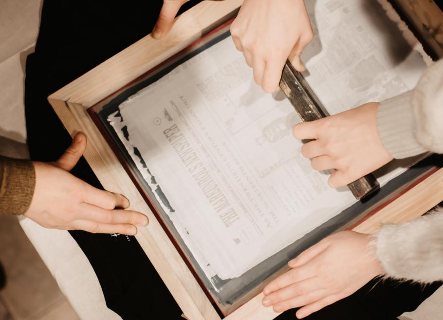 Rokas veido papīru, izmantojot koka rāmi un sietspiedes tehniku, redzams laikraksta fragments zem ūdens.