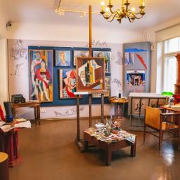 A colorful artist’s studio with paintings, an easel, a desk, and antique furniture.